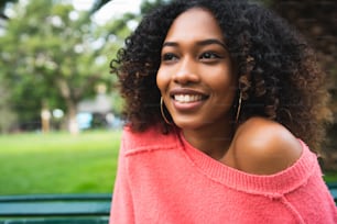 Retrato da jovem mulher afro-americana bonita sentada no banco no parque. Ao ar livre.