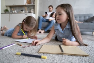 Children sisters playing drawing together on floor while young parents relaxing at home on sofa, little girls having fun, friendship between siblings, family leisure time in living room