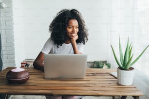 Focused African American female with curly hair sitting at table with humidifier and potted plant and browsing laptop in cozy room at home