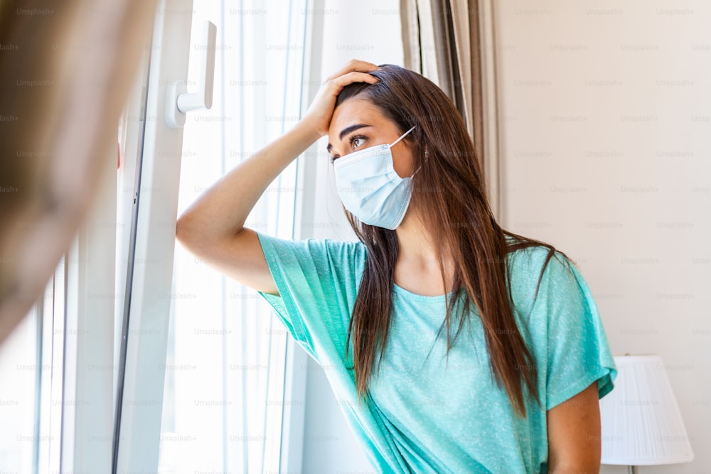 Junge Frau mit Schutzmaske schaut mit Traurigkeit aus dem Fenster, Selbstisolation aufgrund der globalen COVID-19-Coronavirus-Pandemie