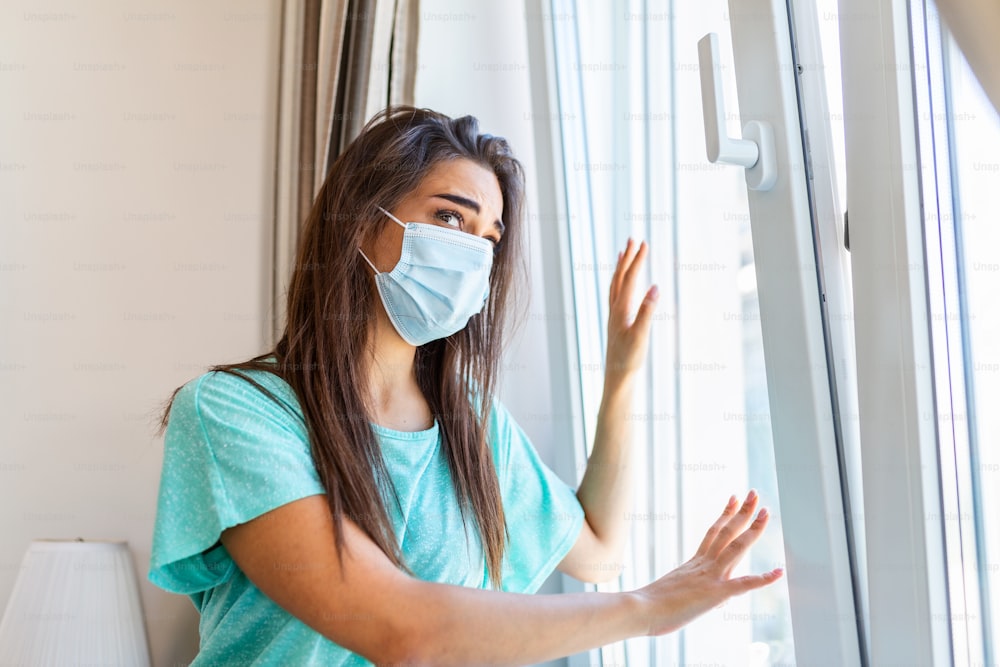 자택 격리. 백인 여성은 의료용 마스크를 쓰고 창문에 앉아 밖을 내다보며 외출하고 싶어합니다. 코로나 바이러스 감염, 전염병, 질병 발생 및 전염병으로부터 보호합니다.