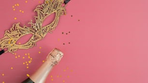 Golden masquerade mask, champagne bottle and confetti on pink background with copy space. Flat lay, top view. Christmas party concept.