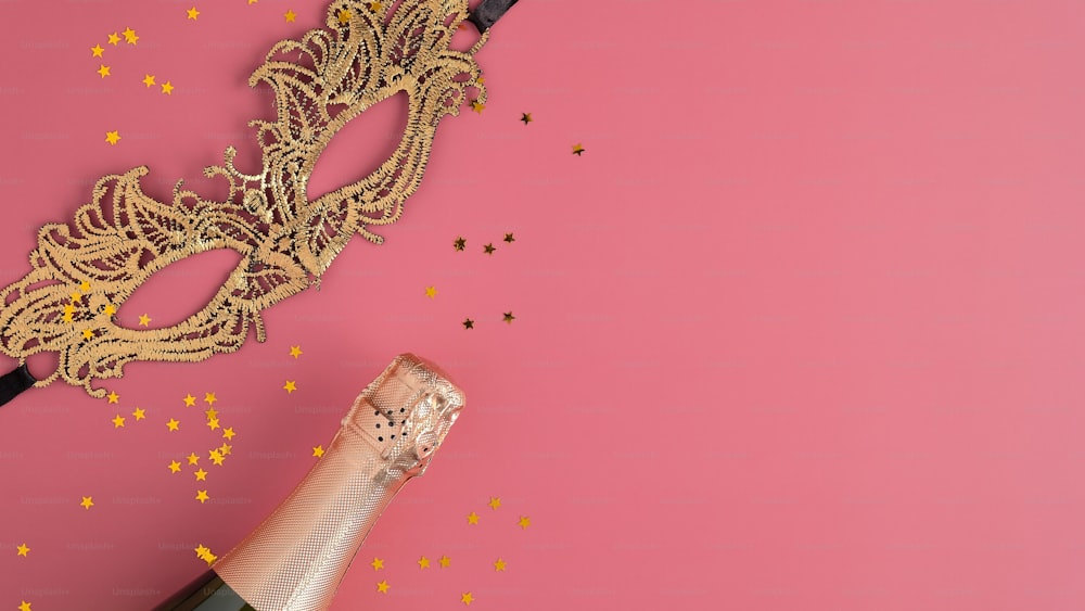 Golden masquerade mask, champagne bottle and confetti on pink background with copy space. Flat lay, top view. Christmas party concept.
