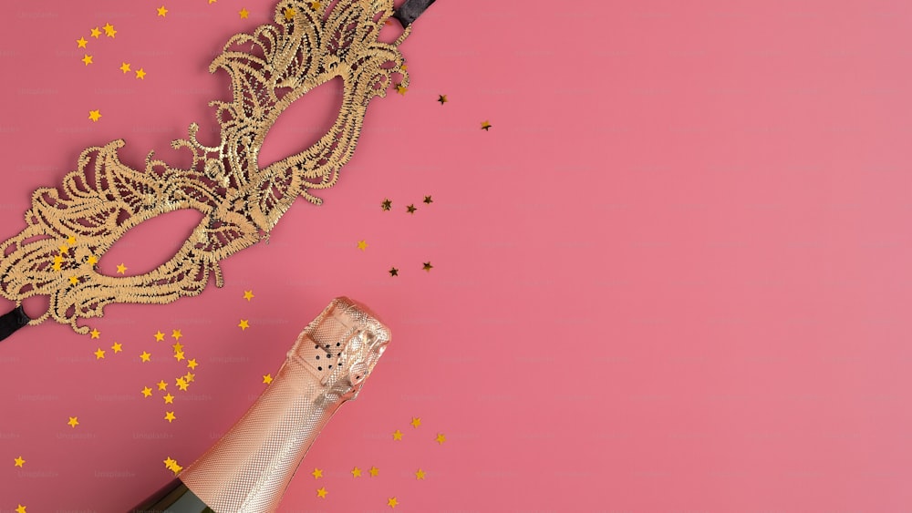 Máscara de mascarada dorada, botella de champán y confeti sobre fondo rosa con espacio de copia. Plano, vista superior. Concepto de fiesta de Navidad.