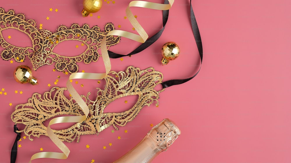 Christmas party masquerade concept. Champagne bottle, golden carnival masks, balls, confetti on pink background. Flat lay, top view.