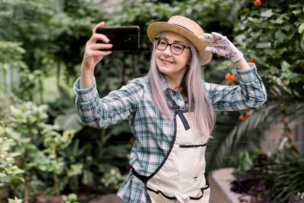 긴 회색 머리를 가진 매력적인 쾌활한 60년대 여성 정원사의 초상화, 체크 무늬 셔츠, 앞치마, 밀짚모자를 쓰고 아름다운 오렌지에 서서 스마트폰으로 셀카를 만들면서 즐거운 시간을 보내고 있습니다.
