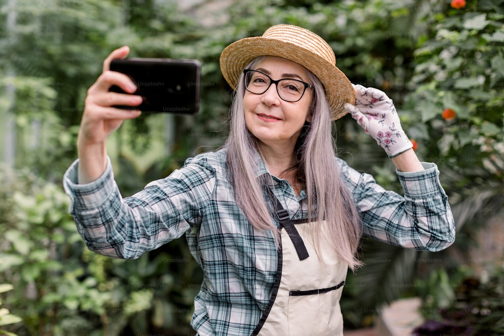 Portrait of attractive cheerful 60s lady gardener with long grey hair, wearing checkered shirt, apron and straw hat, standing in beautiful orangery and having fun while making selfie on smartphone.