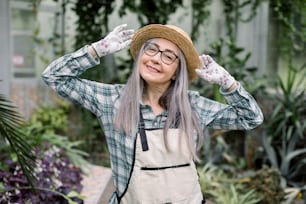 Portrait of gorgeous joyful mature woman gardener with long grey straight hair, wearing checkered casual shirt and apron, smiling to camera with hands on her straw hat, posing in greenhouse.