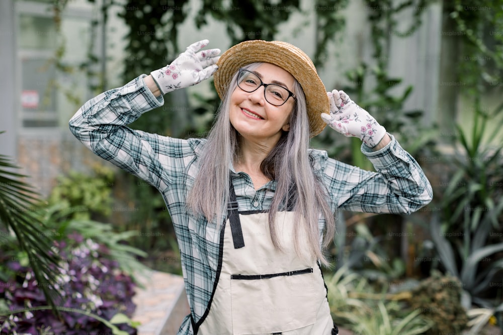 長い灰色のストレートヘアでゴージャスな楽しい成熟した女性の庭師の肖像画は、チェック柄のカジュアルなシャツとエプロンを着て、麦わら帽子に手をかけてカメラに微笑み、温室でポーズをとっています。