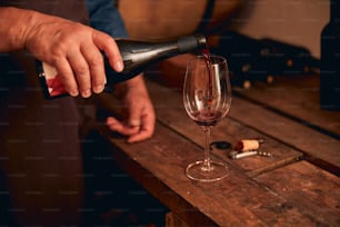 Gros plan d’une main masculine versant une boisson alcoolisée dans un verre à vin tandis qu’un homme se tient près d’une table en bois dans une cave à vin