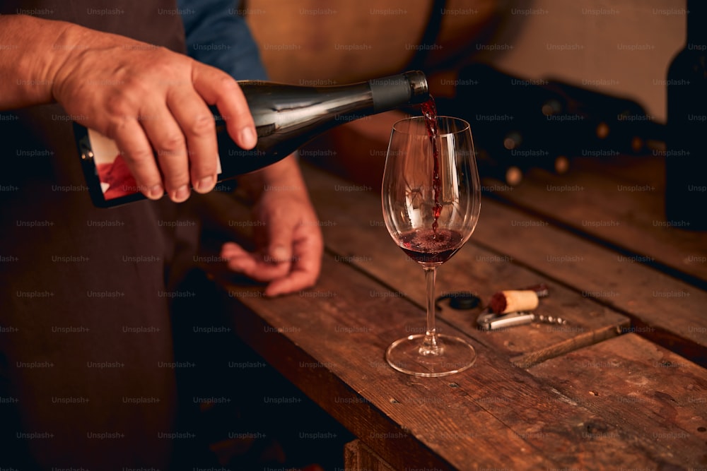 Primer plano de la mano masculina que vierte la bebida alcohólica en la copa de vino mientras el hombre está de pie junto a la mesa de madera en la bodega