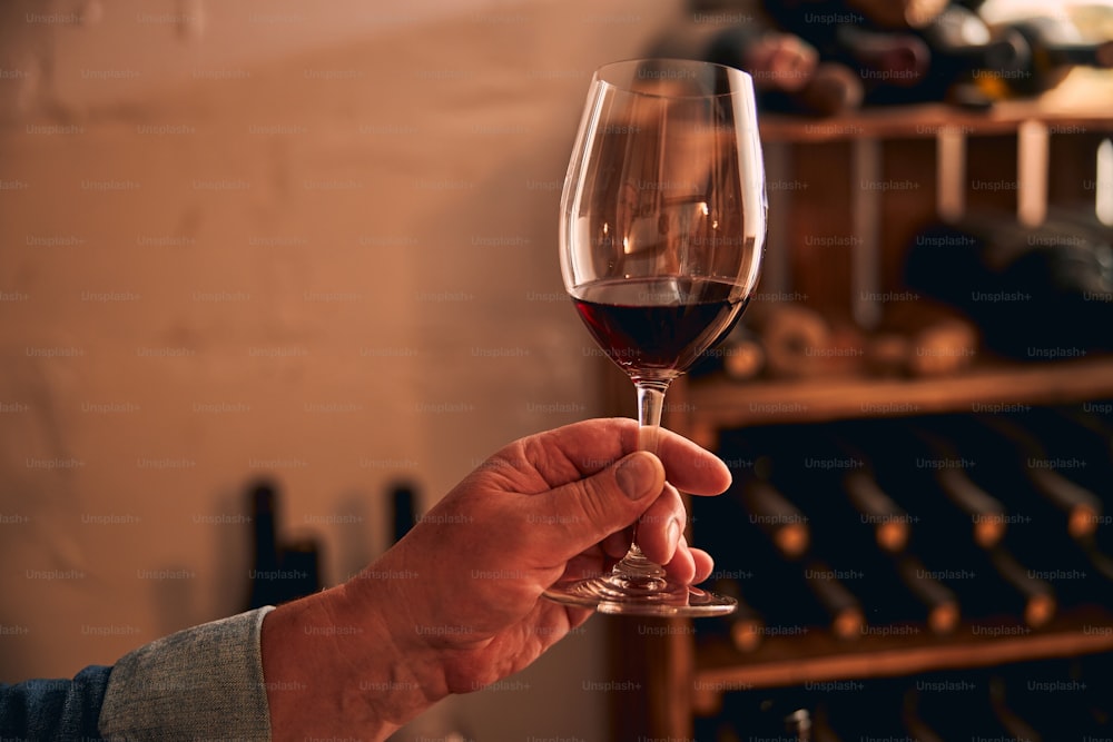 Close up da mão do homem com copo de bebida alcoólica e prateleiras de vinho no fundo desfocado