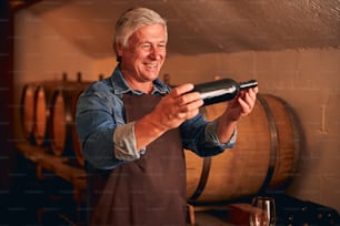 Fröhlicher Winzer in Schürze, der eine Flasche alkoholisches Getränk hält und lächelt, während er Zeit im Weinlager verbringt