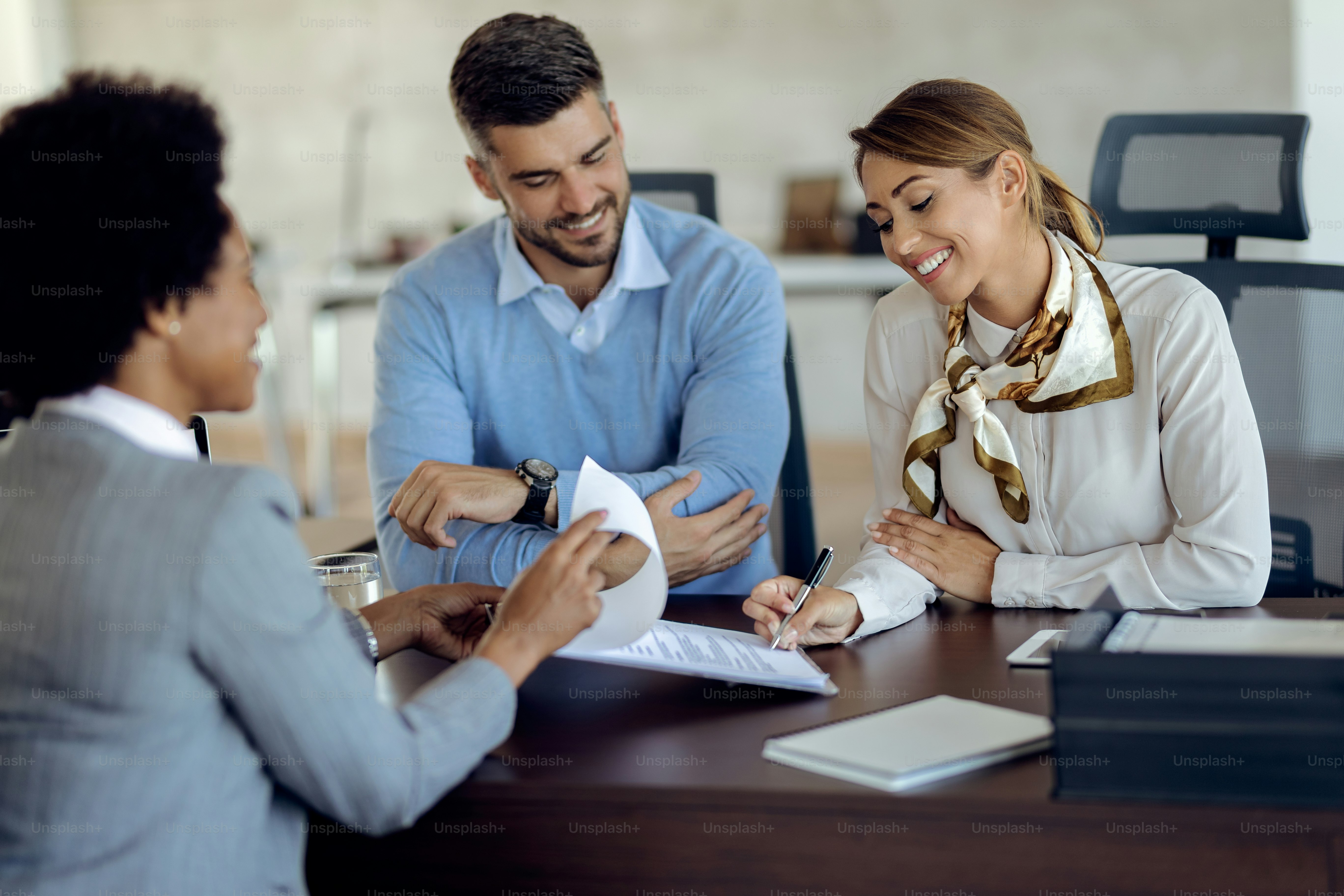 Young happy woman signing a contract while being with her husband on a meeting with insurance agent.