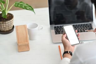 オフィスの木製のテーブルの上に、真っ白な画面とラップトップを備えたモックアップ携帯電話を保持して使用しているトリミングされたショットの女性。