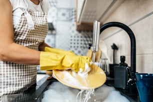 Young adult woman with yellow protective gloves washing her dishes on kitchen sink. Household and home hygiene routine.