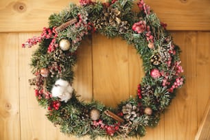 Corona de Navidad que cuelga en la puerta de madera rústica de la casa. Corona navideña tradicional con frutos rojos y adornos, piñas y canela sobre fondo de madera, decoración navideña. Espacio para el texto