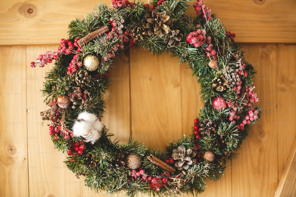 Weihnachtskranz hängt an rustikaler Holztür im Haus. Traditioneller Weihnachtskranz mit roten Beeren und Ornamenten, Tannenzapfen und Zimt auf Holzhintergrund, Urlaubsdekor. Platz für Text