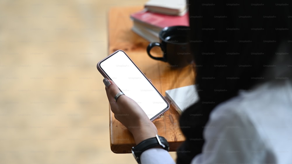 Foto cortada fêmea segurando smartphone com tela vazia na mesa do escritório.