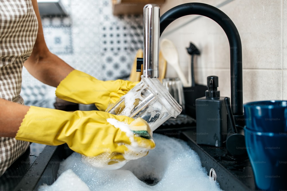 Jeune femme adulte avec des gants de protection jaunes lavant sa vaisselle sur l’évier de la cuisine. Routine d’hygiène domestique et domestique.