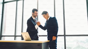 Two happy business people celebrate at office meeting room. Successful businessman congratulate project success with colleague at modern workplace while having conversation on financial data report.
