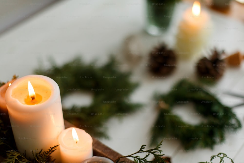 Burning candles on rustic background with christmas wreath, pine cones and ornaments on wooden table in evening. Holiday workshop advent, cozy atmosphere. Copy space