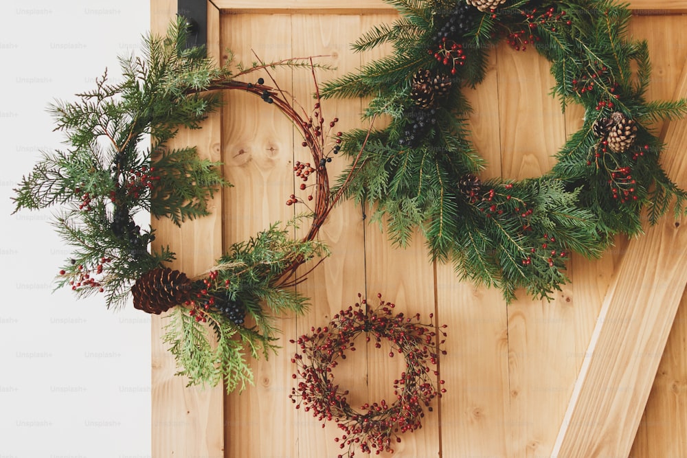 Coronas navideñas creativas naturales y diferentes que cuelgan en una elegante puerta rural. Feliz Navidad. Coronas rústicas de Navidad en puerta de madera en el interior, decoración festiva de vacaciones
