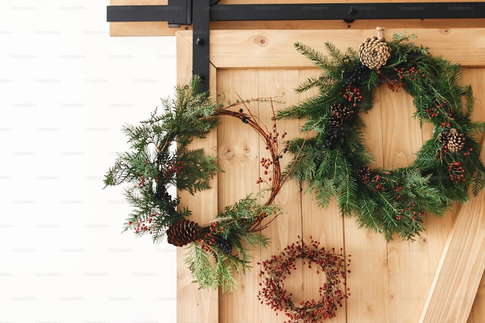 Coronas navideñas creativas naturales y diferentes que cuelgan en una elegante puerta rural. Feliz Navidad. Coronas rústicas de Navidad en puerta de madera en el interior, decoración festiva de vacaciones