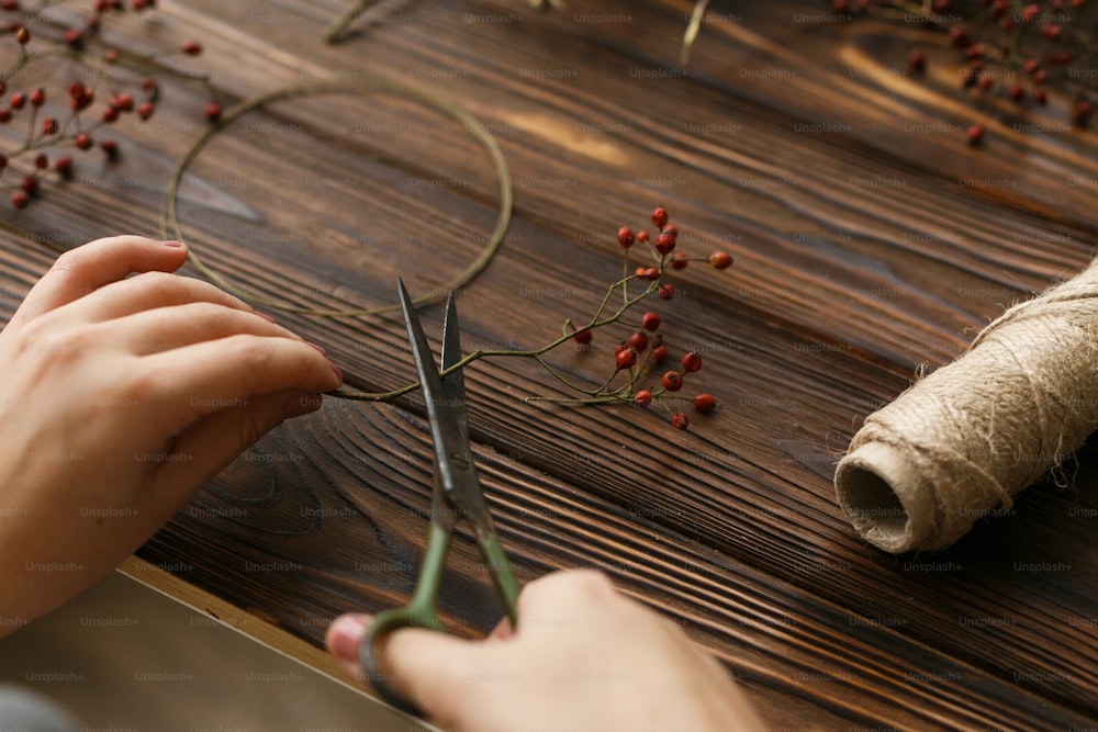Making christmas wreath on rustic wooden table, holiday advent. Florist making simple christmas wreath with red berries, holding scissors. Festive workshop