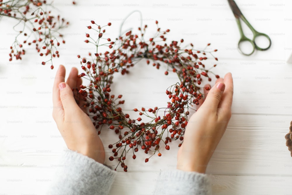 Making christmas wreath on white wooden table at home, holiday advent. Female hands making simple christmas wreath with red berries on white wooden table with thread and scissors