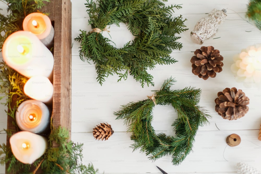 Rustic christmas wreath with candles, pine cones, scissors and ornaments on white wooden table, flat lay. Making simple stylish christmas wreath with fir and cedar branches, holiday advent