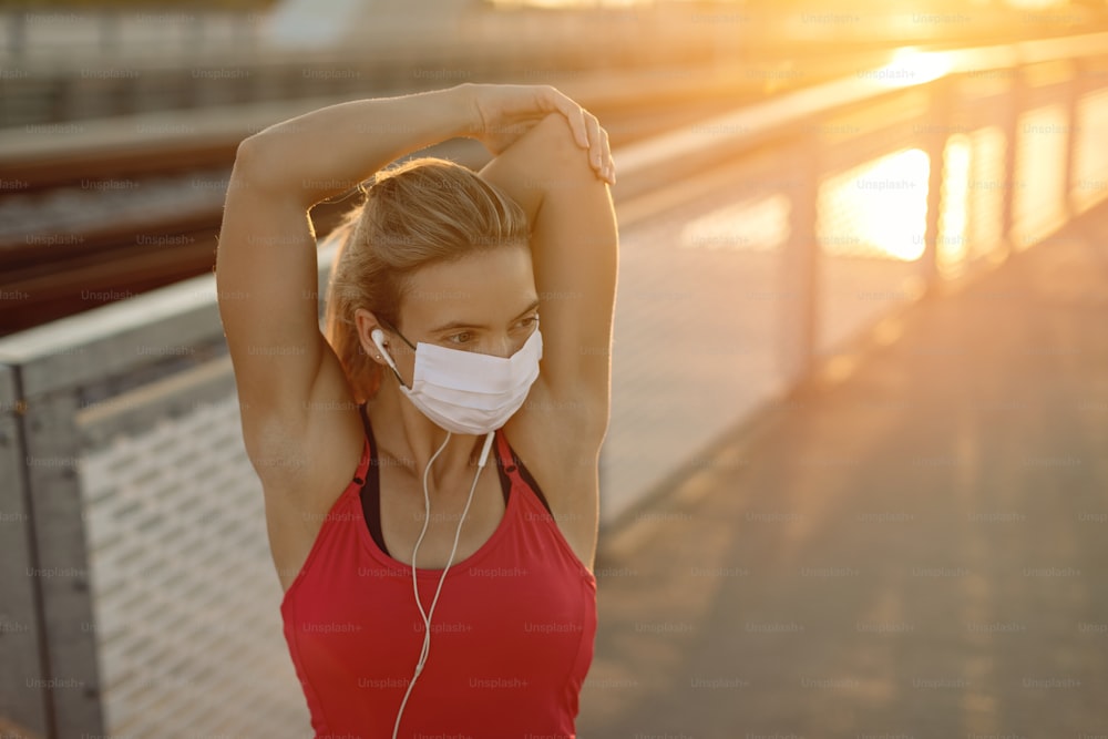 Atleta femenina con máscara facial protectora mientras hace ejercicios de estiramiento al aire libre al atardecer. Espacio de copia.