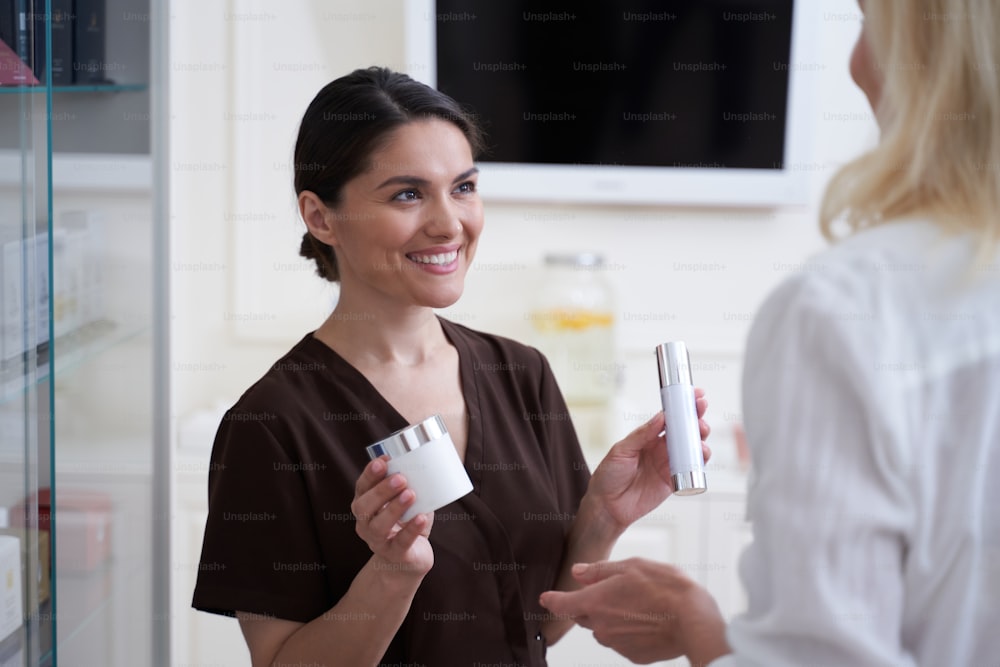 Happy beautician standing in beauty clinic and showing professional cosmetics for skin care for woman