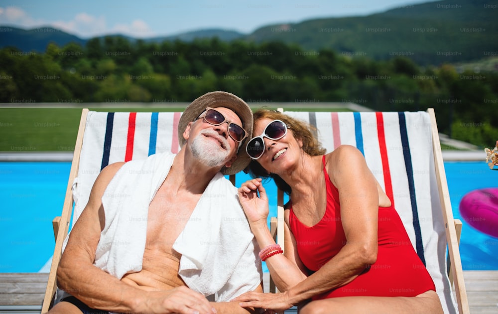 Cheerful senior couple sitting by swimming pool outdoors in backyard, looking at camera.