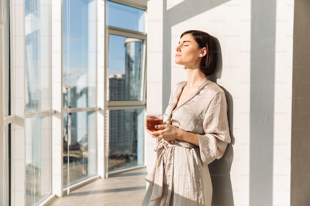 Elegant woman with short dark hair in housecoat holding glass of tea and enjoying sunny weather while standing near window in posh apartment