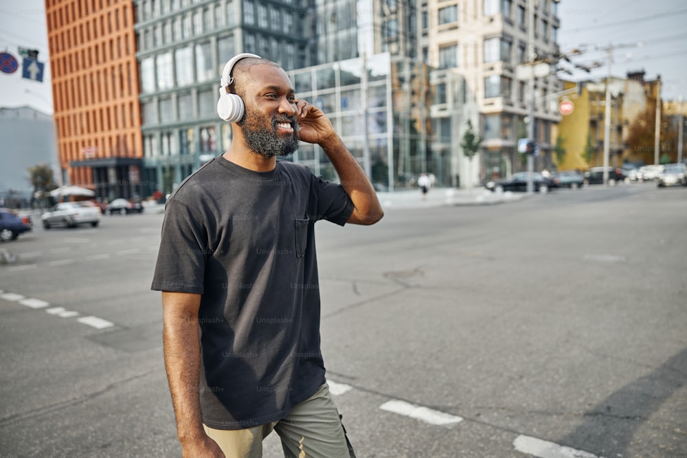 Contented young man in casual clothes walking in the city center and touching the wireless headphones on his head. Website banner