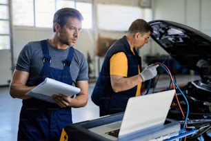 Two mechanic examining vehicle breakdown in auto repair shop. Focus is on mechanic using laptop while writing notes on clipboard.