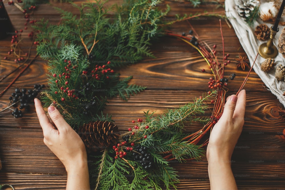 Couronne de Noël rustique moderne sur table en bois, vacances de l’Avent. Mains féminines tenant une couronne de Noël élégante avec des baies rouges, des branches de cèdre vert et une pomme de pin. Confection d’une décoration festive