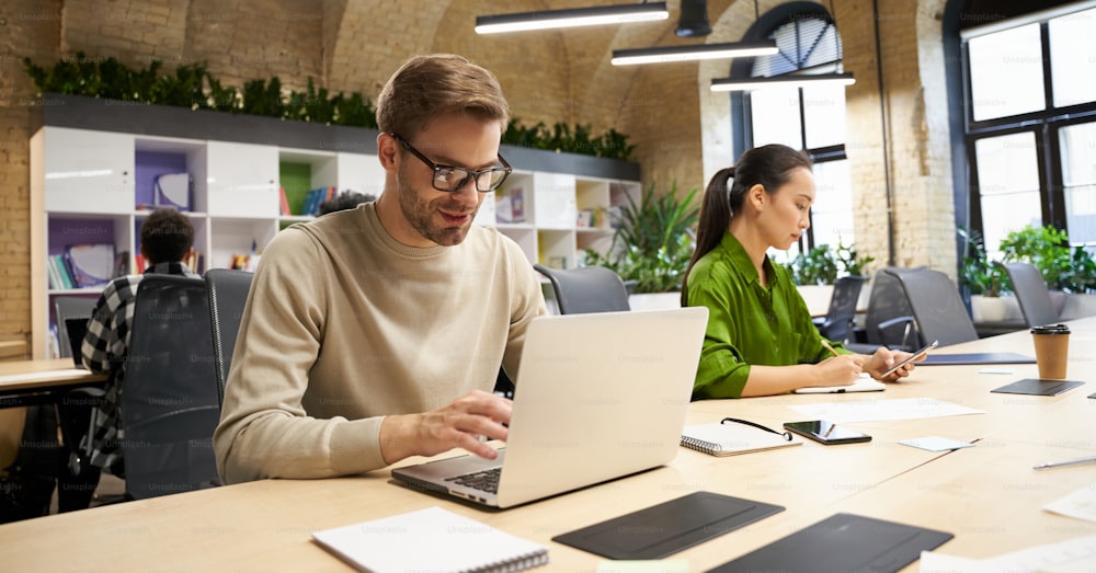 Young focused caucasian man wearing eyeglasses working on laptop while sitting at desk with his female colleague in the modern coworking space. Business people and teamwork concept