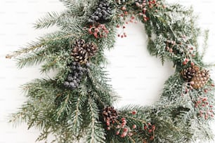 Rustic christmas wreath hanging on white wall, festive modern decoration. Creative stylish christmas wreath with red berries and fir branches close up, isolated in white. Merry Christmas