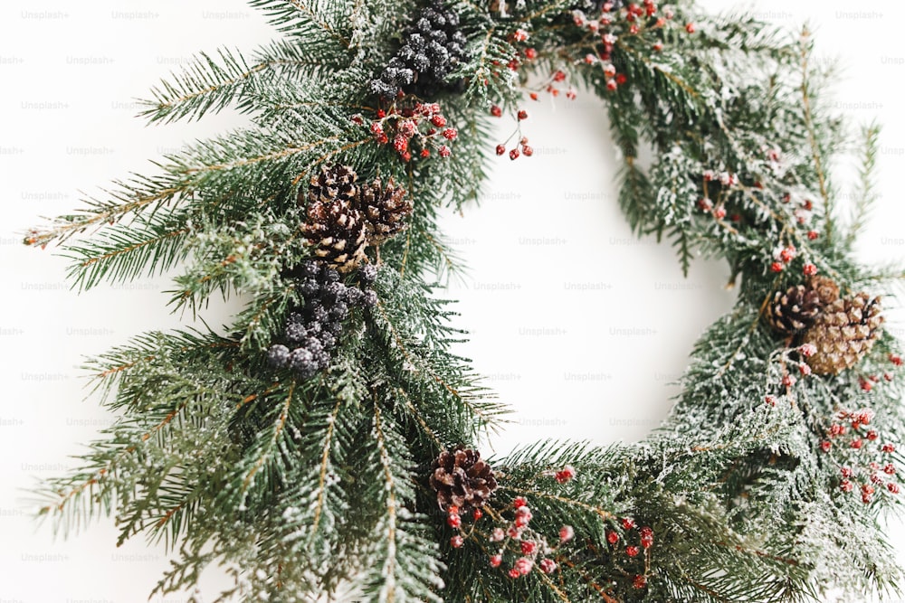 Ghirlanda di Natale rustica appesa alla parete bianca, decorazione moderna festiva. Ghirlanda di Natale elegante creativa con bacche rosse e rami di abete da vicino, isolata in bianco. Buon Natale