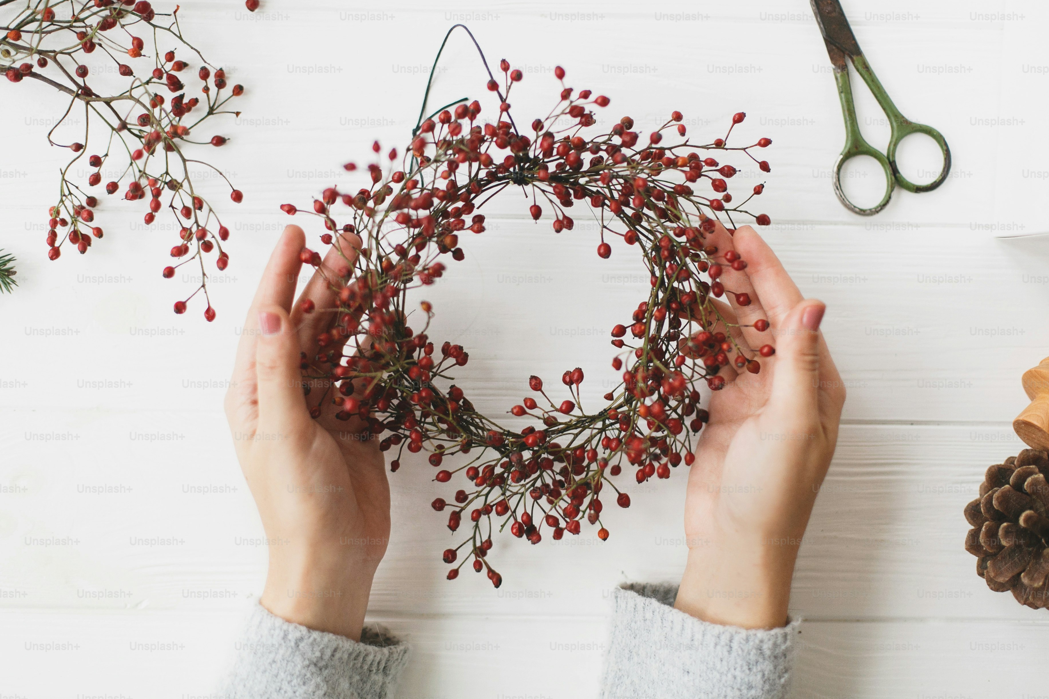 Making christmas wreath on white wooden table flat lay, holiday advent. Female hands holding simple christmas wreath with red berries on white wooden table with thread and scissors