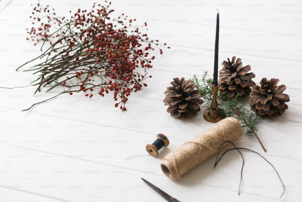 Détails pour couronne de Noël rustique sur table en bois blanc. Baies rouges, fil, ciseaux, pomme de pin et bougie sur fond rustique,
Faire une couronne de Noël à la maison, l’Avent des vacances