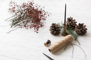 Details for rustic christmas wreath on white wooden table. Red berries, thread, scissors, pine cone and candle on rustic background,
making christmas wreath at home, holiday advent