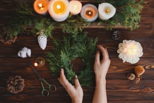 Corona navideña rústica con velas, piñas, tijeras y adornos sobre mesa de madera blanca, plana. Manos de florista que hacen una corona navideña elegante simple con ramas de abeto, adviento de vacaciones