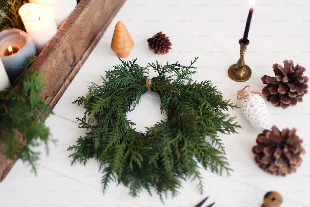 Rustic christmas wreath with candles, pine cones, thread and ornaments on white wooden table. Making simple stylish christmas wreath with cedar branches, holiday workshop advent