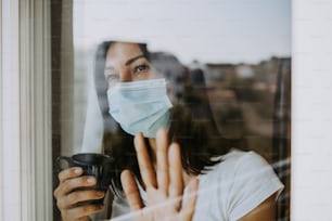 A frightened and worried woman looks out the window of her apartment or hospital room. She wears a protective mask on her face for fear of a dangerous and contagious virus. Quarantine and social distancing concept.