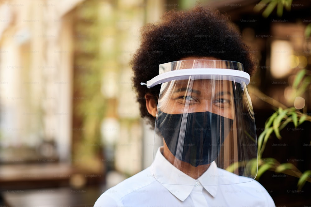 Camarera afroamericana feliz con máscara facial protectora y visera mientras trabaja en un café al aire libre durante la epidemia de coronavirus.