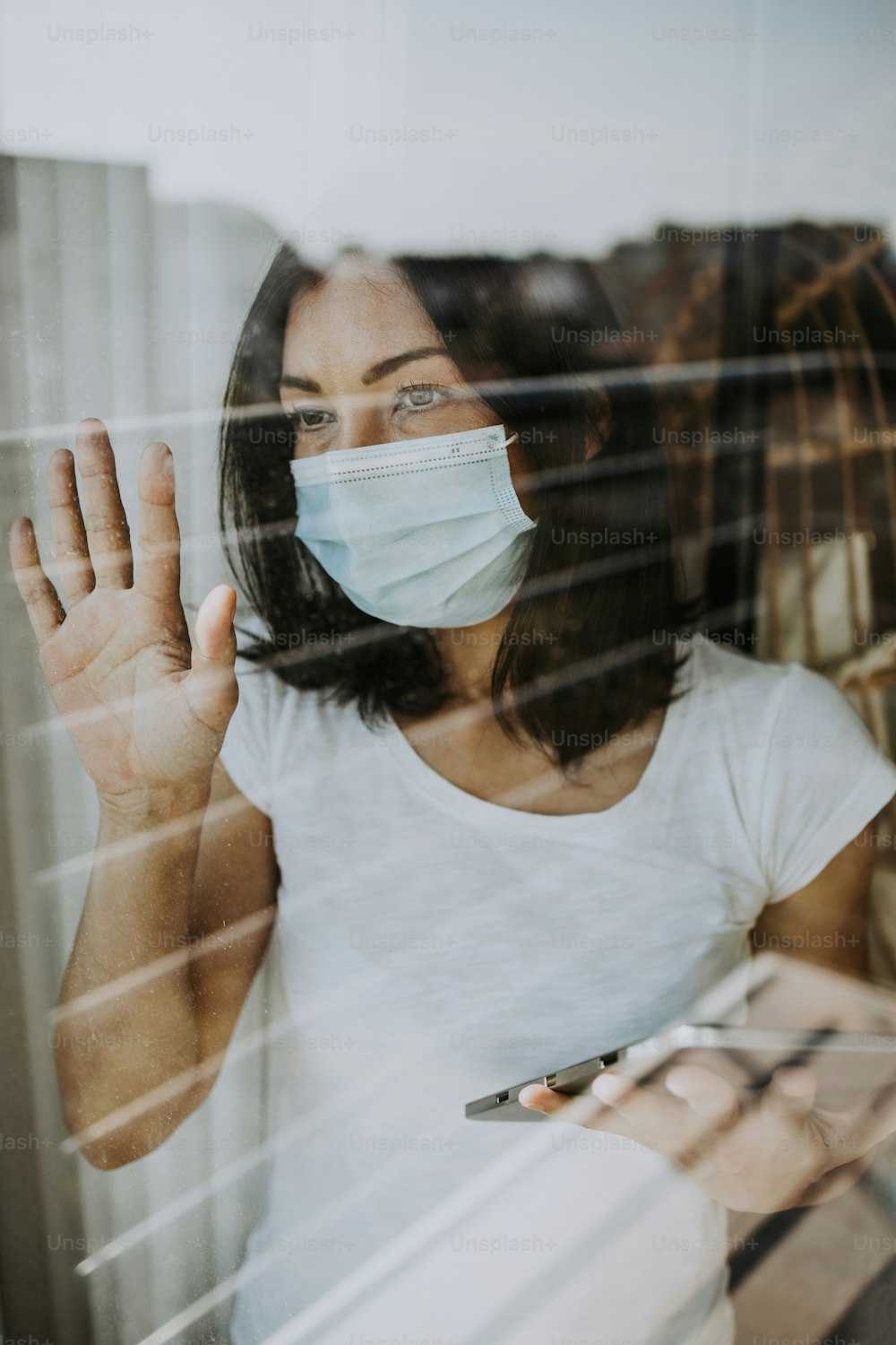 A frightened and worried woman looks out the window of her apartment or hospital room. She wears a protective mask on her face for fear of a dangerous and contagious virus. Quarantine and social distancing concept.