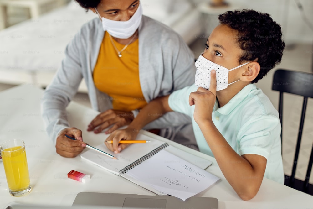 African American little boy wearing protective face mask and thinking while doing homework with his mother.